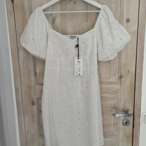 Studentklänning - Superfin vit kortklänning från Holly & Whyte by Lindex med broderade blommor, puffärmar och smock i ryggen. Klänningen har v-ringning och dragkedja i sidan. är i storlek M och passar perfekt till student, sommaren och finare tillfällen.