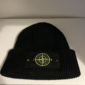 Stone Island Mössa - Svart ribbstickad mössa från Stone Island med klassisk uppvikt kant och ikonisk logopatch framtill. Mössan har en snygg, enkel design och är tillverkad i mjukt stickat material som håller dig varm. Perfekt för dig som vill ha en clean streetwear-look.