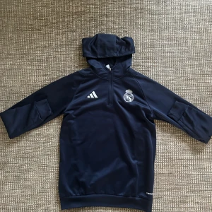 Mörkblå Real Madrid hoodie Adidas - Snygg mörkblå hoodie från Adidas med Real Madrid-logga på bröstet och klassiska ränder i vitt, grått och gult på ärmarna. Tröjan har huva, lång ärm och half zip framtill. Perfekt för fotbollsträning eller chill dagar.