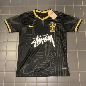 Svart Brasil x Stüssy Nike fotbollströja - Exklusiv svart Brasil fotbollströja från Nike i samarbete med Stüssy. Tröjan har gula detaljer på krage och ärmslut, Brasil-logga på bröstet och Stüssy-tryck framtill. Tillverkad i Dri-FIT-material som andas, perfekt för fotboll eller streetwear.