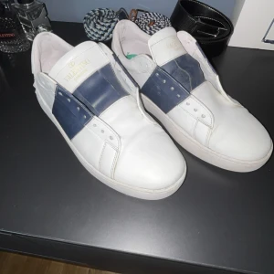 Valentino Garavani open  - (SKO SNÖREN KOMMER OCKSÅ TILL) Snygga vita sneakers från Valentino Garavani med bred marinblå rem över vristen. Skorna har rund tå, platt sula och är tillverkade i skinn. Perfekta för dig som gillar stilrena och exklusiva sneakers med en unik touch.