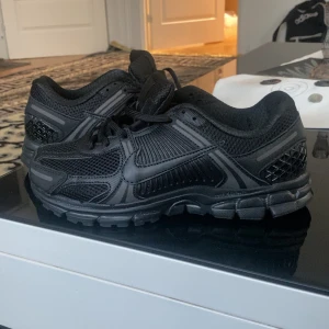 Nike Vomero 5 - Snygga helsvarta Nike sneakers med coola detaljer i mesh och syntet. Skorna har en chunky sula, snörning och diskret Swoosh-logga på sidan. Perfekta för dig som gillar sportig och stilren vibe.