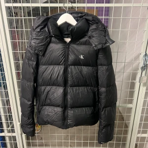 Svart pufferjacka från Calvin Klein Jeans - Svart pufferjacka från Calvin Klein Jeans med huva och dragkedja framtill. Jackan har ett stilrent CK-logo på bröstet och är tillverkad av återvunnen nylon och polyester med dunfyllning. Vattenavvisande och perfekt för kalla dagar.