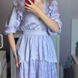 Ljuslila volangklänning från Maje - Superfin ljuslila klänning från Maje med broderade vita detaljer, volanger på ärmar och kjol samt v-ringning. Klänningen har trekvartsärmar och markerad midja. Använd en gång på halloween förra året! 