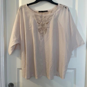 Beige blus med spets från Zara Basic - Säljer en ljusbeige blus från Zara Basic med vackra spetsdetaljer och broderier framtill vid halsringningen. Blusen har korta, lösa ärmar och en avslappnad passform. Perfekt för dig som gillar romantiska och feminina detaljer.