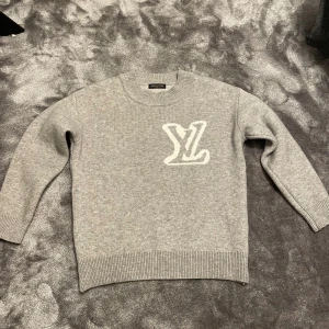 Grå stickad Louis Vuitton tröja - Supersnygg grå stickad tröja från Louis Vuitton med stort vitt LV-logo på bröstet. Rund hals och ribbade muddar vid ärmslut och nederkant. Perfekt för dig som gillar exklusiva märken och vill ha en clean look.