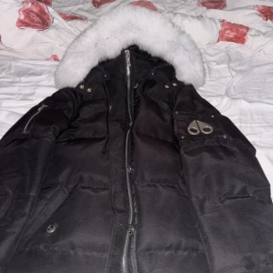 Svart Moose Knuckles dunjacka med päls - Säljer en svart dunjacka från Moose Knuckles med vit päls på huvan och ikonisk logga på ärmen. Jackan har dragkedja framtill, flera fickor med knappar och detaljer i metall. Perfekt för kalla dagar och har en riktigt snygg streetstil.