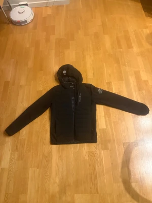 Svart pufferjacka från Sail Racing - Svart ”cardigan” som är vind- och vattentät från Sail Racing med huva och snygga logodetaljer på både bröst och rygg. Jackan har dragkedja framtill och är quiltad över kroppen. Perfekt för dig som vill ha en stilren och sportig look under kyliga dagar.
