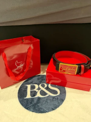 Louboutin black and red belt 110cm - Bilderna visar ett bälte från Christian Louboutin med röd insida och guldfärgat spänne, men ingen väst syns. 110cm