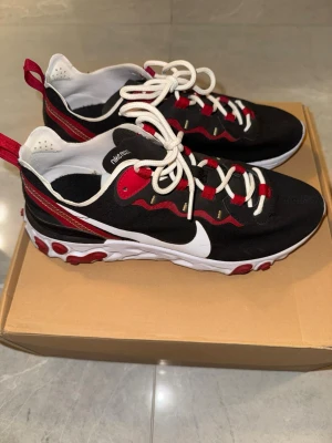 Nike React Element 55 svart/röd sneakers - Nike React Element 55 sneakers i svart med röda och vita detaljer. Snygg vågig design på sidan, vit sula med röda gummipluppar undertill och stora vita swoosh-loggor. Ovandel i syntetmaterial, vita snören och röda dragflikar bak. Perfekt för dig som gillar sportig och modern stil.