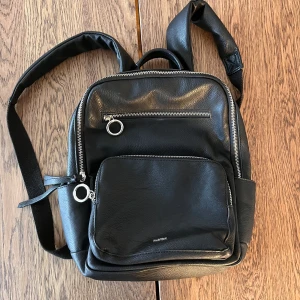 Liten ryggsäck - Ryggsäck med silvriga detaljer. Använd, men den har inga särskilda defekter annat än normalt slitage och ett skav på undersidan (hör av dig om du vill ha bild på det). Den är ca 30x28x12 cm i storlek.