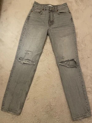 Grå ripped jeans med raka ben - Snygga ljusgrå jeans med raka ben och slitna detaljer över knäna. Klassisk femficksmodell med hög midja och dragkedja. Perfekta för en avslappnad streetstil och tillverkade i robust denim.