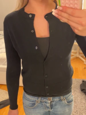 Svart kofta  - Svart, stilren kofta från BOSS i superfine merinoull. Klassisk rund hals och knäppning framtill med svarta knappar. Snygg passform och långärmad modell som funkar perfekt till jeans eller kjol. Mjuk och tunn kvalitet som känns lyxig mot huden.