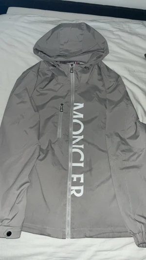 Grå vindjacka från Moncler - Snygg grå vindjacka från Moncler med stor logga längs dragkedjan. Storlek M passar även L. Jätte bra skick. Pris kan diskuteras!