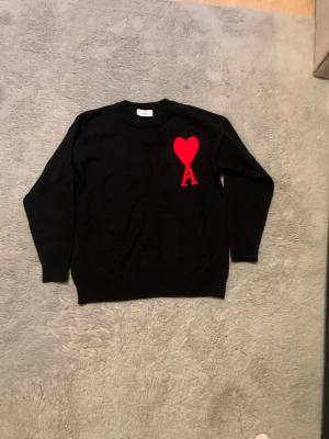 Svart stickad tröja från Ami - Svart stickad tröja från Ami med röd hjärt- och A-logga på bröstet. Rund hals, ribbade muddar och klassisk passform. Materialet är mjuk bomull, perfekt för att ge din outfit en clean och modern vibe. Pris 800 storlek S 