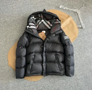 Svart Burberry dunjacka med avtagbara ärmar - Svart dunjacka från Burberry med avtagbara ärmar och huva. Insidan har klassiskt rutigt Burberry-mönster i grått, vitt och rött. Jackan har dragkedja, knappar och två fickor med dragkedja. Perfekt för dig som vill ha både jacka och väst i ett.