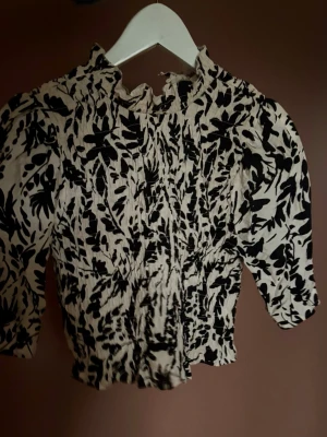 Beige och svart blommig blus - Säljer en beige blus med svart blommigt mönster och smockad struktur. Blusen har hög hals med volangkant och en lätt croppad passform. 