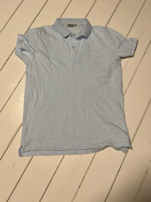 Ljusblå piké från RYVLS - Säljer en stilren ljusblå pikétröja från H&M med klassisk krage och knappar framtill. Perfekt för dig som gillar enkel och clean stil. Kortärmad och i mjukt bomullsmaterial som känns skönt mot huden.