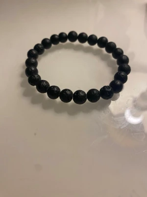 Svart pärlarmband med lava-sten - Stilrent svart armband med runda pärlor av lava-sten. Pärlorna har en matt, lite grov yta som ger en cool och unik känsla. Perfekt accessoar för dig som gillar minimalistisk stil och vill ha något som sticker ut på handleden.