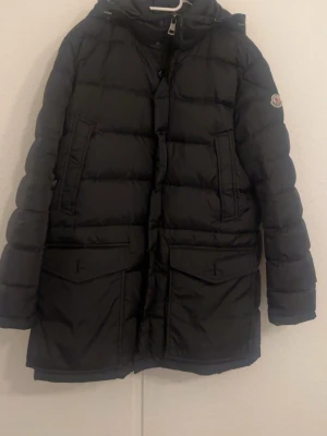 Moncler cluny - Säljer en svart Moncler dunjacka med snygg pälsdetalj på huvan. Jackan har långa ärmar, flera fickor med lock och dragkedja framtill. Klassisk Moncler-logga på ärmen och coolt serietryck på insidan. Perfekt för kalla dagar och riktigt stilren.
