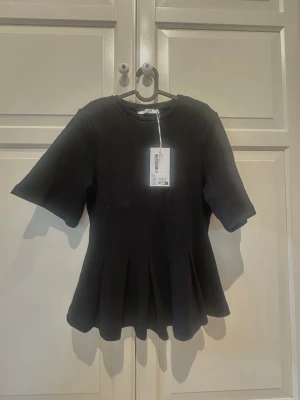 Svart blus med plissering från NA-KD - Svart blus från NA-KD med rund halsringning och korta, vida ärmar. Toppen har en utsvängd midja med veck. Oanvänd med prislappen kvar. 