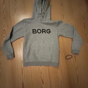 Grå hoodie från Björn Borg - Snygg grå hoodie från Björn Borg med svart BORG-tryck på bröstet. Tröjan har huva, långa ärmar och en klassisk känguruficka framtill. Tillverkad i mjuk bomullsmix som är skön att ha på sig hela dagen.