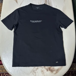 Svart t-shirt med snakeprint från Smog - Svart t-shirt från Smog i relaxed fit med vit text och tryck. Framsidan har ett litet citat på bröstet, medan ryggen har stort tryck med två ormar, taggar och texten 'Outlaw Apparel'. Tillverkad i mjuk bomull och har klassisk rund hals.