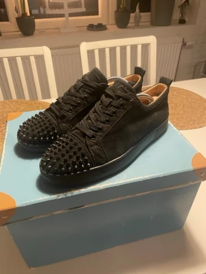 Svarta Christian Louboutin sneakers med nitar - Svarta sneakers från Christian Louboutin med coola svarta nitar på tån och klassisk röd sula. Skorna har snörning och är tillverkade i mocka med läderdetaljer på insidan. Snygg och edgy design som sticker ut, perfekt för dig som gillar statement pieces. (Kan skicka bild på mer)