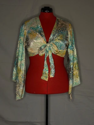 Färgglad omlott bolerotopp i satin - Unik bolerotopp i glansigt satinmaterial med turkosa, gula och beige toner. Toppen har långa vida ärmar, omlottmodell och knyt framtill. Mönstret är en mix av paisley och geometriska former som ger en cool boho-vibe.