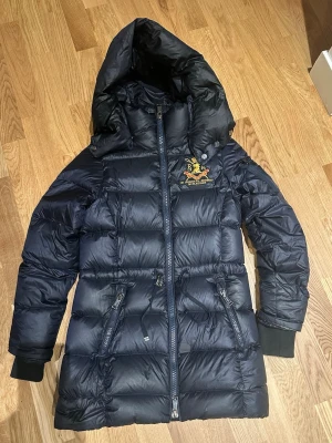 Mörkblå dunjacka Polo Ralph Lauren XS - Snygg mörkblå dunjacka från Polo Ralph Lauren med broderad emblem på bröstet, avtagbar huva och två dragkedjeförsedda fickor. Jackan har snörning i midjan och ribbade muddar vid ärmslut. Perfekt för kalla dagar och har en stilren, lång siluett. Obs den har en patch där en cigarett kom på den när jag var och gick på stan