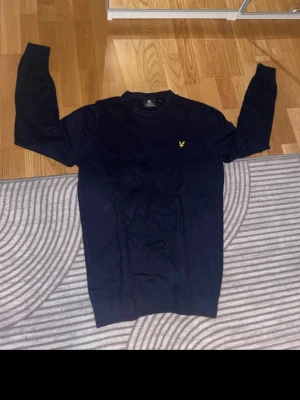 Mörkblå stickad tröja Lyle & Scott - Snygg mörkblå stickad tröja från Lyle & Scott med klassisk gul logga på bröstet. Rund hals och långa ärmar, perfekt för dig som gillar stilrena och enkla plagg. Materialet är mjukt och skönt, passar bra till jeans eller chinos.