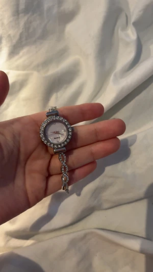 Glittrig silverfärgad klocka - Säljer en elegant klocka i silverfärg med glittrande stenar runt urtavlan och på länken. Urtavlan är rund och har en ljus ton, och armbandet har snygga detaljer med infällda stenar. Perfekt accessoar för att lyfta din outfit.