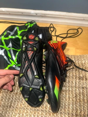 Adidas svarta och orange fotbollsskor - Säljer ett par Adidas fotbollsskor med snygg design i svart, orange och grönt. Skorna har snörning och mönstrad ovansida med Adidas-logga på hälen. Sulan har gröna dobbar för bra grepp på planen. Materialet är syntet och skon har en rund tå.