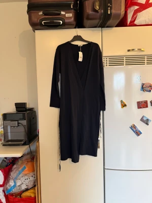 Mörkblå omlottklänning från Filippa K - Snygg mörkblå klänning från Filippa K i mjuk jersey. Klänningen har långa ärmar, v-ringning och knytband i midjan för en stilren och enkel look. Perfekt för dig som gillar minimalistisk stil och vill ha något bekvämt men ändå elegant. Storlek M. Helt ny med tags/ prislapp fortfarande kvar. 
