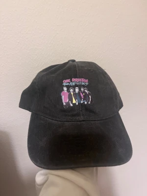 One Direction svart keps On The Road Again - Svart keps med justerbart spänne bak och tryck framtill med One Direction och texten 'On The Road Again Tour 2015'. Motiv med bandmedlemmarna i färgglada kläder. Perfekt för fans som vill visa sin stil.