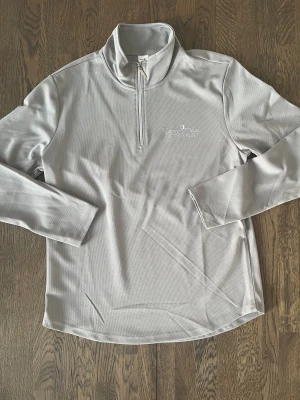 Ljusgrå golftröja Next Tee - Snygg ljusgrå långärmad tröja från Next Tee med half zip och hög krage. Materialet är polyester med en diskret textur och logga broderad på bröstet. Perfekt för golf eller chill dagar. Enkel och stilren design som funkar till många outfits.