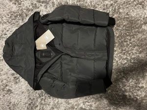 Svart pufferjacka från Canada Goose - Svart pufferjacka från Canada Goose med huva och snyggt märke på ärmen. Jackan har dragkedja och knappar framtill, ribbade muddar vid ärmslut och quiltad design. Perfekt för kalla dagar och har en clean, modern look.