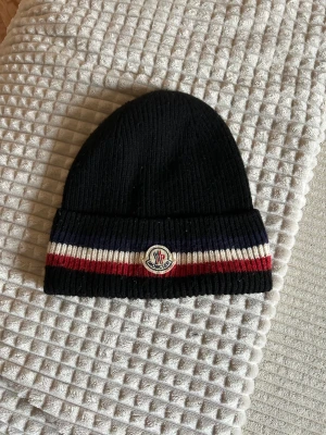 Svart Moncler mössa med ränder - Snygg svart ribbstickad mössa från Moncler med bred uppvikt kant och klassiska ränder i marinblått, vitt och rött. Märkets logga sitter framtill på kanten. Perfekt accessoar för att hålla värmen och addera lite lyx till din look.
