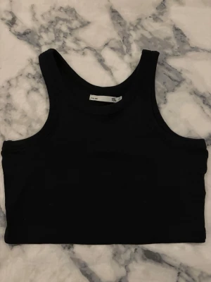Svart croppad linnetopp från 157 - Svart croppad linnetopp från 157 i storlek XS. Toppen har en klassisk rund halsringning och är ärmlös med breda axelband. Materialet är ribbad bomull som ger en skön och stretchig passform. Perfekt för dig som gillar enkel och stilren design.