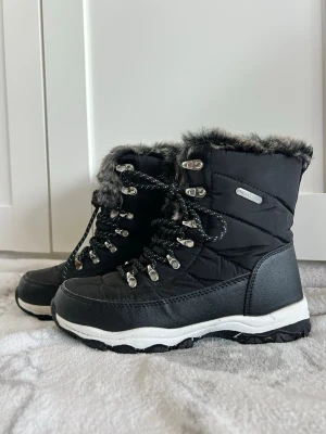 Svarta vinterboots med fuskpäls - Säljer ett par svarta vinterboots från Winterproof med fuskpäls runt skaftet och snörning framtill. Bootsens ovandel är quiltad och har detaljer i syntet och skinnimitation. Sulan är grov och vit med svart mönster för extra grepp.