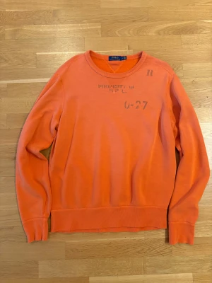 Orange sweatshirt från Polo Ralph Lauren - Snygg orange sweatshirt från Polo Ralph Lauren i storlek L. Tröjan har tryck med text och siffror framtill, rund hals och ribbade muddar vid ärmslut och nederkant. Perfekt för dig som gillar streetstyle och vill sticka ut med färg.
