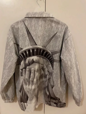Grå overshirt med Frihetsgudinnan-print - Unik grå overshirt med stort konstnärligt print av Frihetsgudinnan som täcker ansiktet med händerna. Jackan har dragkedja framtill, klassisk krage och elastiska muddar vid ärmsluten. Materialet är syntet. Jackan är helt ny också, går ner i pris vid snabb affär