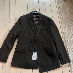 Brun Svart kavaj från Mango Man, stl 48 - Dubbelknäppt brun svart kavaj från Mango Man i klassisk modell. Kavajen har spetsiga slag, fyra fickor och knappar vid ärmslut. Helt ny med prislapp