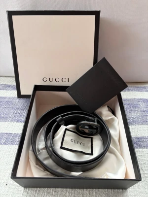 Svart skärp från Gucci i skinn - Säljer ett svart skärp från Gucci i äkta skinn med den ikoniska GG-loggan i svart metall. Bältet är stilrent och passar perfekt till jeans eller kostymbyxor. Kommer med originalbox och dustbag. En riktig klassiker för dig som gillar lyxiga accessoarer. Fullset med kvitto från 2018.