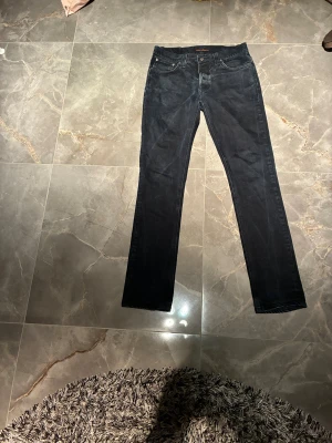 Svarta jeans från Nudie Jeans, 33/36 - Säljer ett par svarta jeans från Nudie Jeans i storlek 33/36. Modellen har klassisk femficksdesign, raka ben och normal passform. Jeansen är tillverkade i mjukt denimtyg och har subtila kontrastsömmar. Perfekt för dig som gillar stilrena och tidlösa jeans.