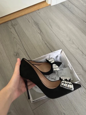 Svarta pumps med strassrosett - Snygga svarta pumps från Seastar med spetsig tå och hög klack. Skorna är i mocka-look och har en stor rosett framtill dekorerad med glittrande strassdetaljer som verkligen sticker ut. Perfekta för dig som vill ha något extra till outfiten.