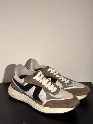 J.lindberg skor - Säljer ett par snygga sneakers i vitt och brunt med svarta detaljer på sidorna. Skorna har en rund tå, platt sula och är tillverkade i en mix av mocka och syntetmaterial. De har klassisk snörning och en sportig siluett som passar till allt.