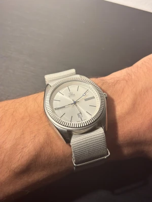 Custom made watch - Bilderna visar en silverfärgad klocka från BOOM Watches med ljusgrått textilband, minimalistisk urtavla och datumvisning. Klockan är fullt fungerande och i nytt skick. 40mm