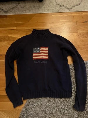 Mörkblå polotröja Ralph Lauren - Snygg mörkblå polotröja från Ralph Lauren med stickad amerikansk flagga på bröstet och broderad logga under. Tröjan har ribbad krage, muddar och nederkant. Perfekt för dig som gillar klassisk stil med en twist.