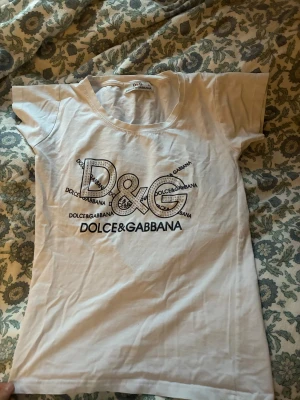 Vit D&G t-shirt med strassdetaljer - Snygg vit t-shirt från Dolce & Gabbana med stort D&G-tryck och strassdetaljer på bröstet. Modellen är kortärmad och har en klassisk rund halsringning. Materialet är mjuk bomull som känns skönt mot huden. Perfekt för dig som gillar statement-plagg och exklusiva märken.